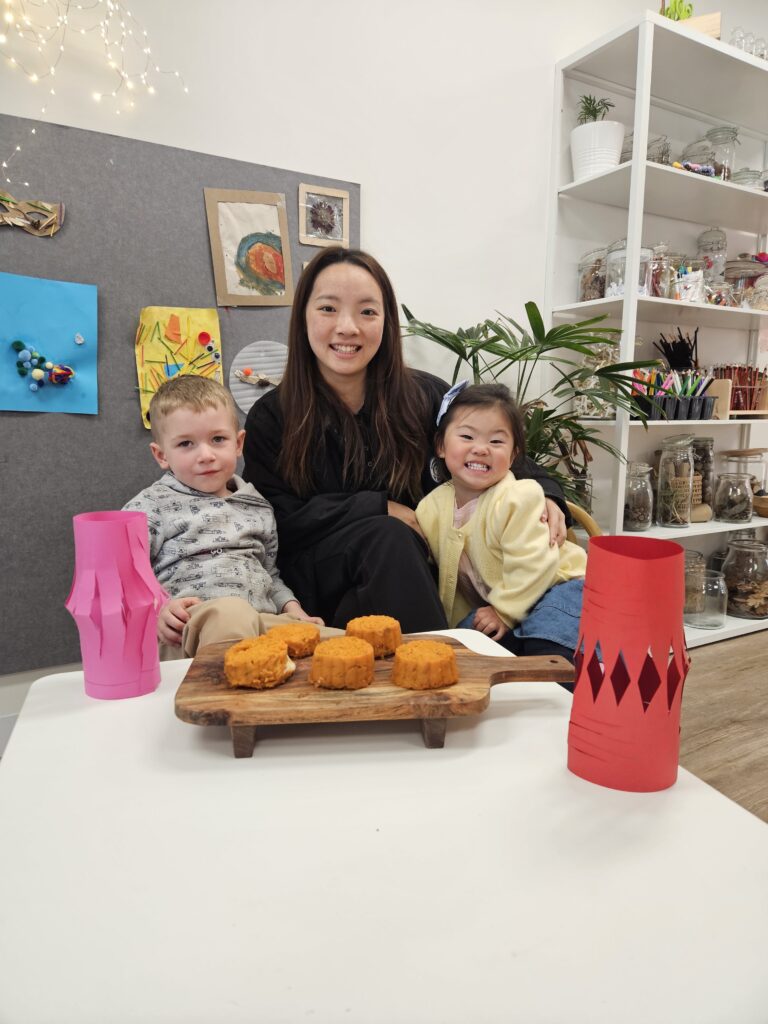 A childcare educator and two children are in front of a table with a mooncake.
